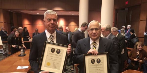 Image of two gentleman standing shoulder-to-shoulder holding up framed pictures